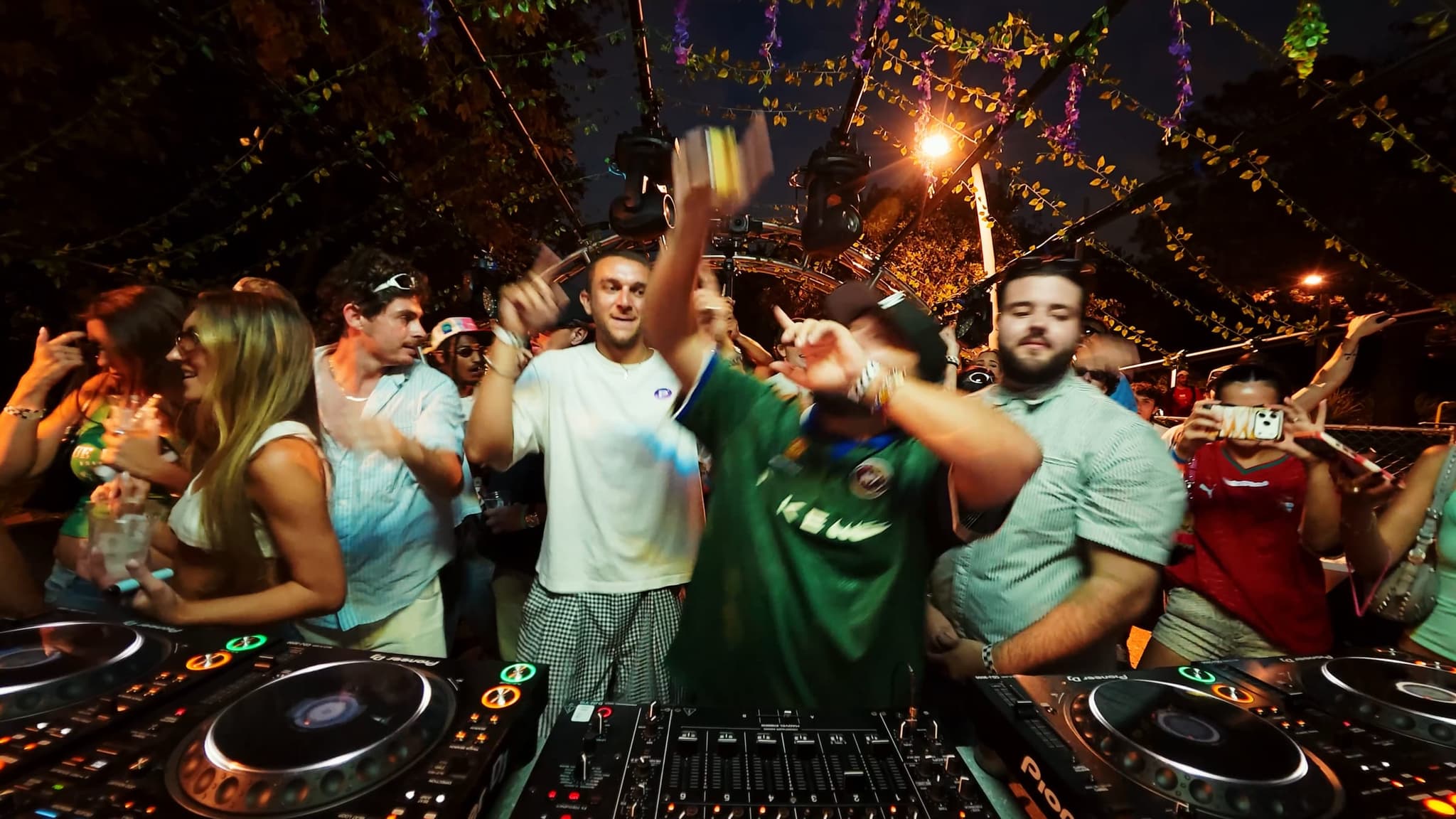 Two DJs with arms in the air behind the decks under the vine-covered arch