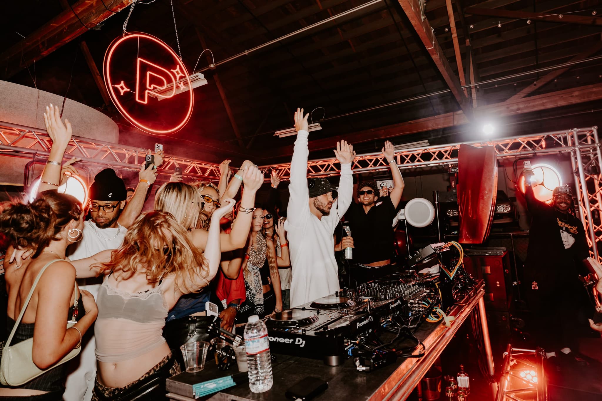 Arms raised around the DJ booth beneath a red neon Product Pluto sign