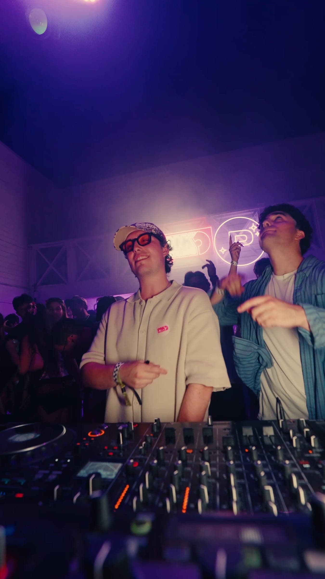 Two DJs in the booth under purple light, the neon emblem behind them