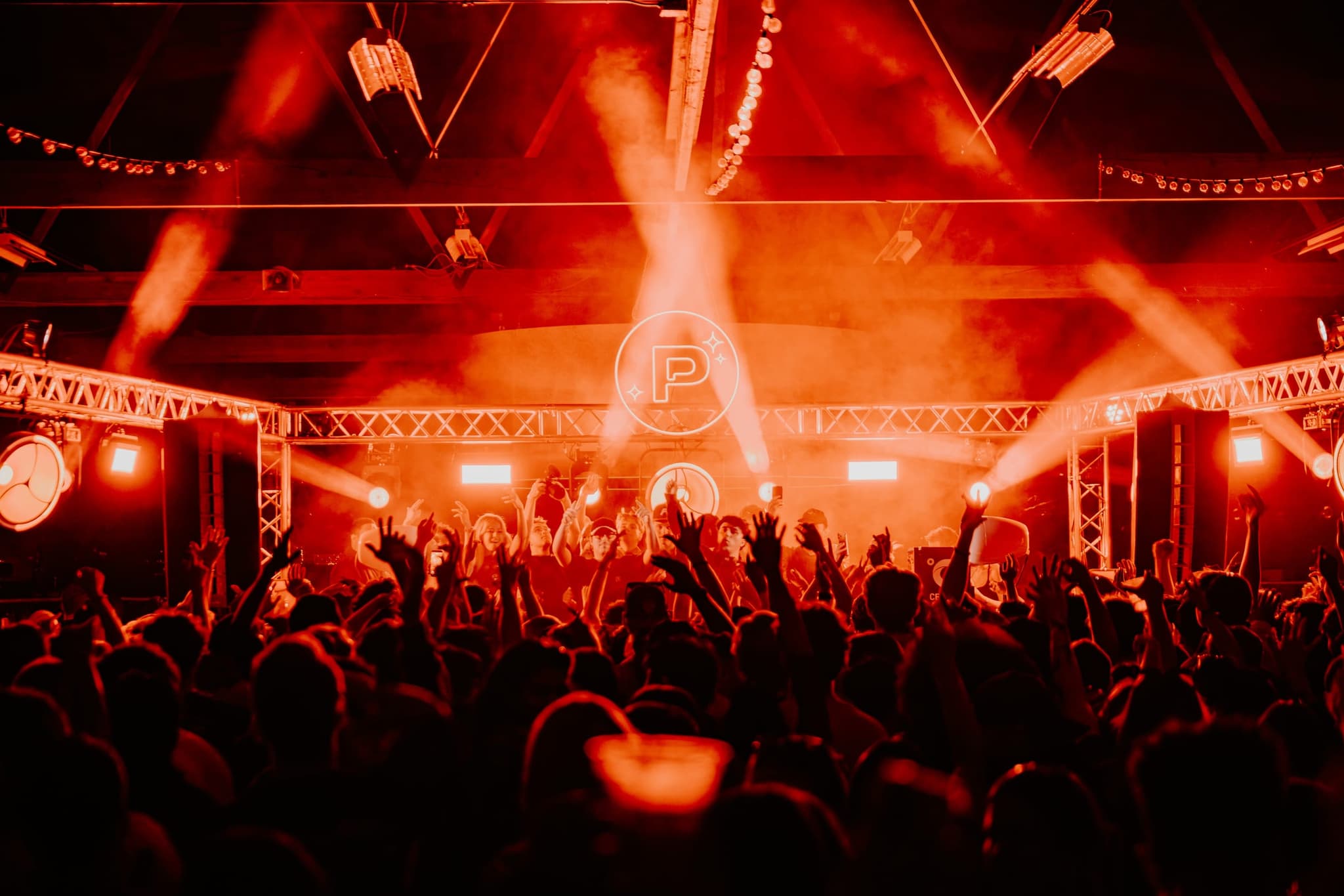 Red light and smoke over a packed warehouse floor, the Product Pluto emblem glowing above the stage