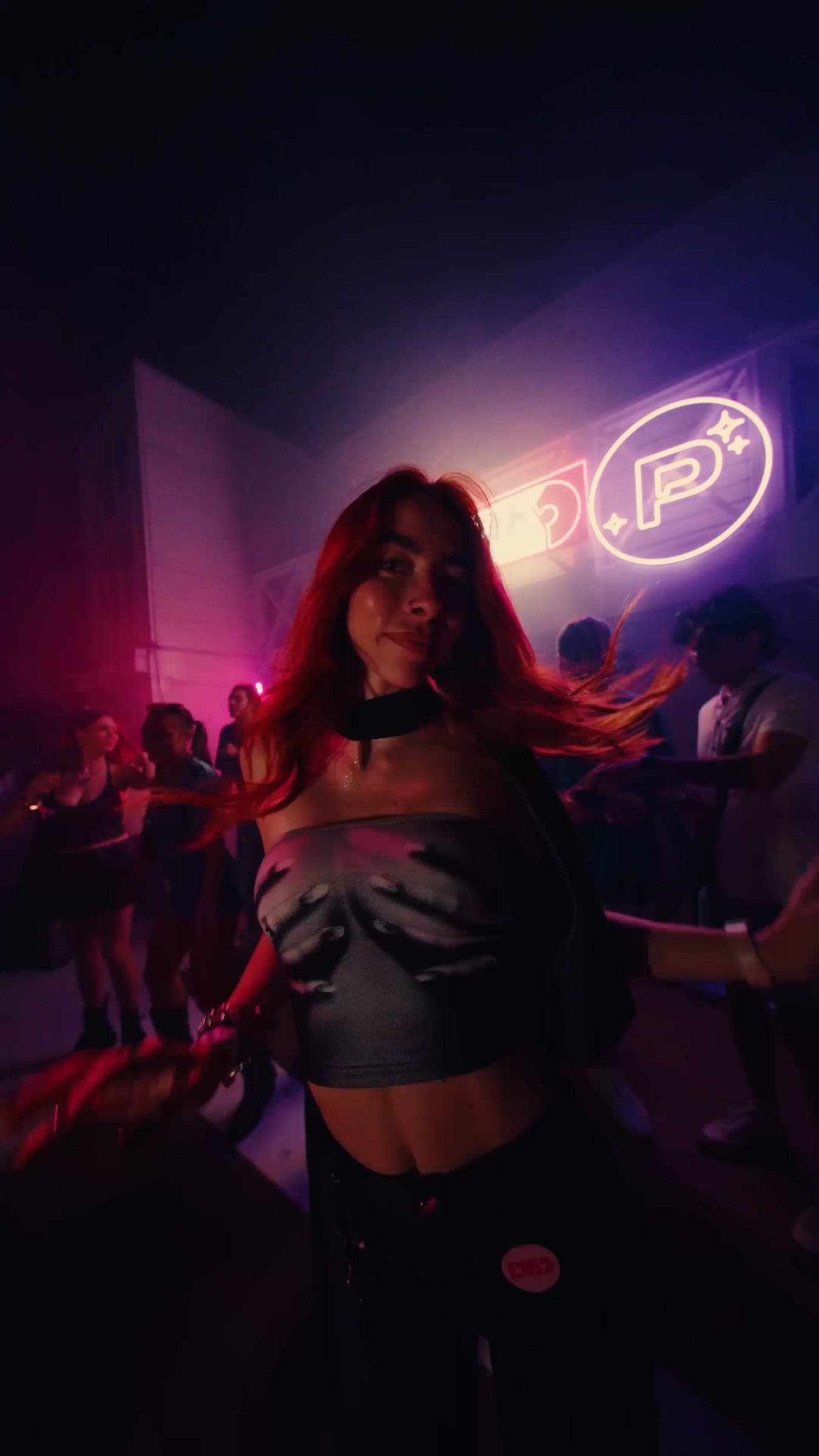 A dancer with red hair under pink light, the neon emblem on the wall behind her