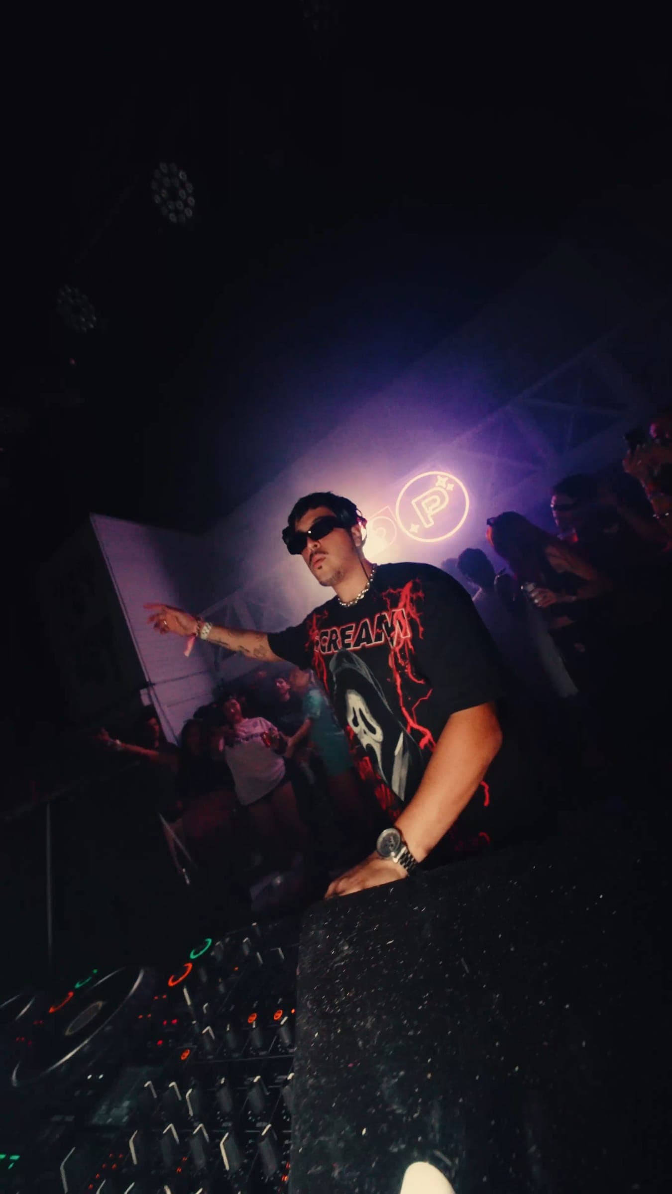 A DJ in sunglasses leaning out from the booth, the neon P sign glowing violet