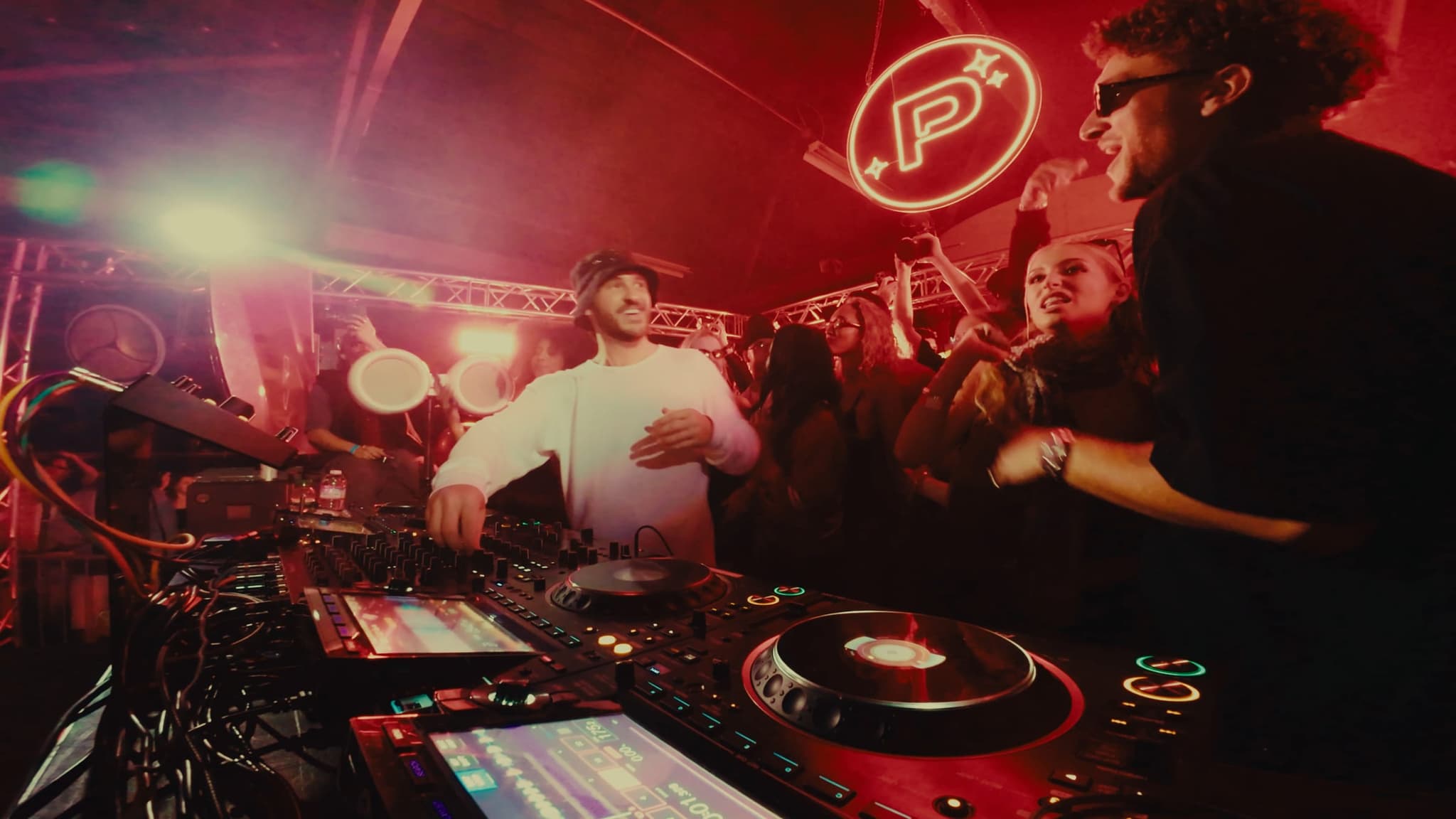 The DJ in white at the decks, the neon emblem glowing red behind him