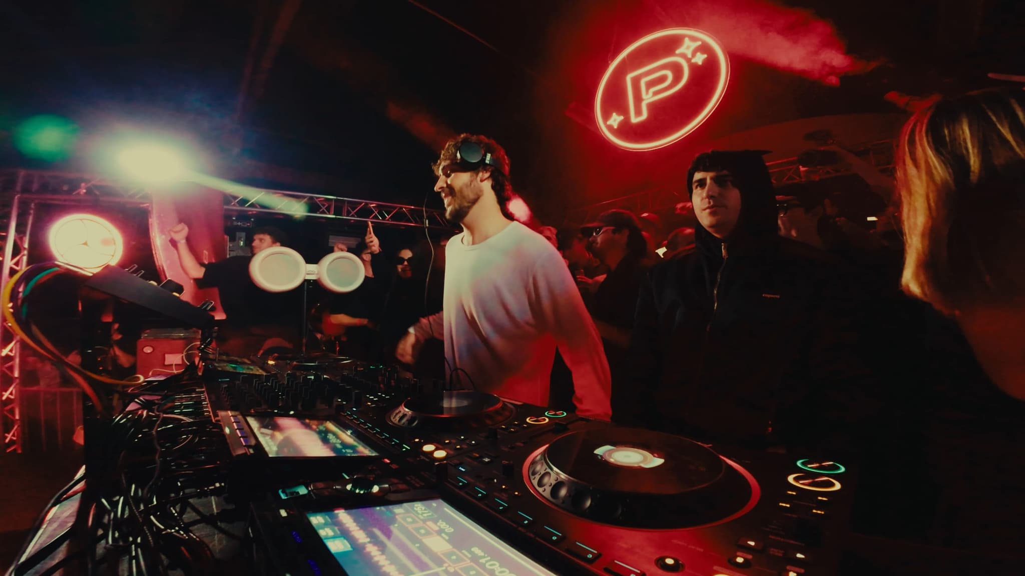 The DJ at the decks with the neon emblem and a green spotlight behind him